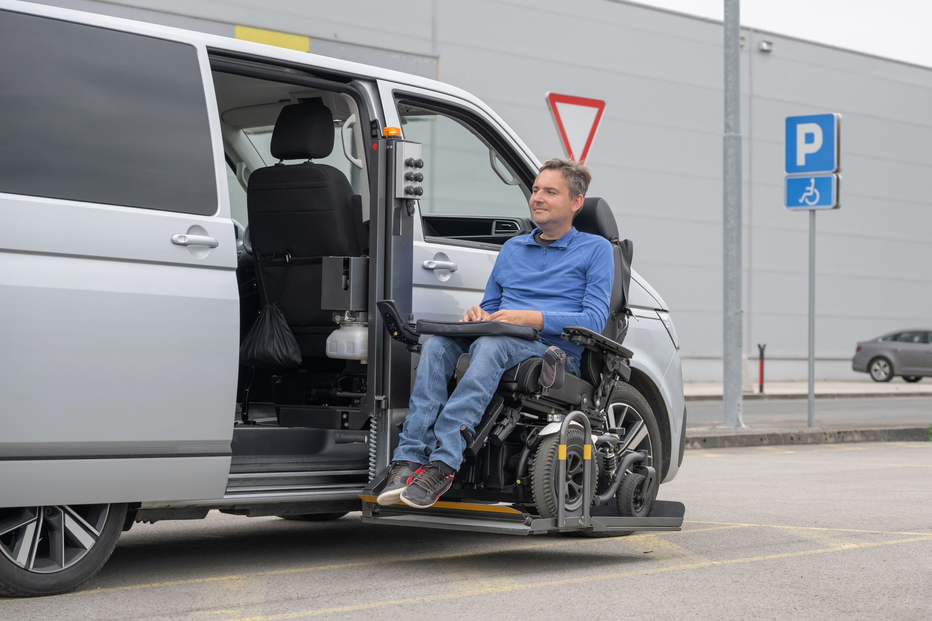 Man in a power wheelchair using a side-entry platform lift to exit a silver wheelchair accessible van in an accessible parking space.