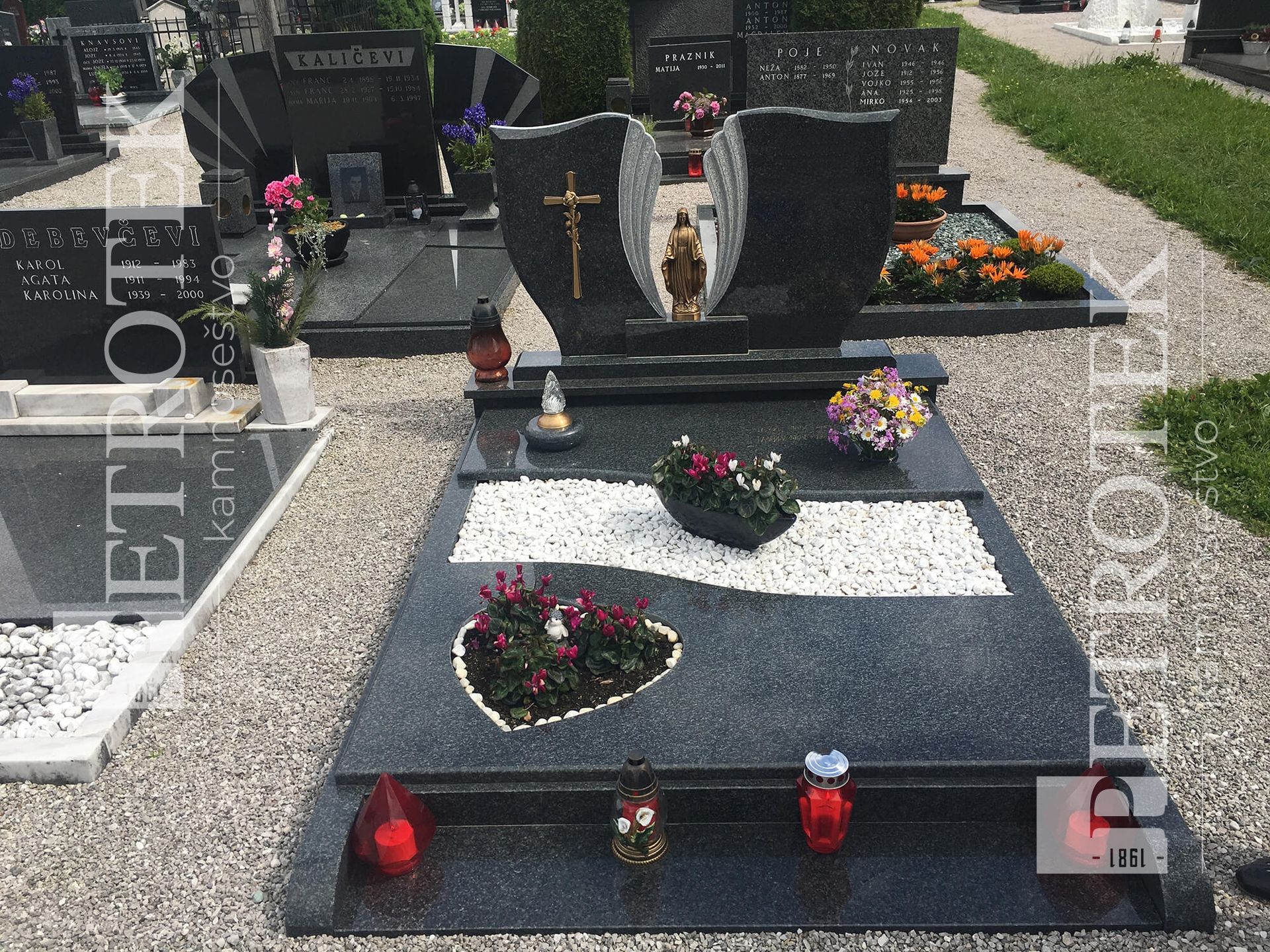 Črn granitni nagrobnik s križem, cvetjem v obliki srca in prižganimi svečami na pokopališču.