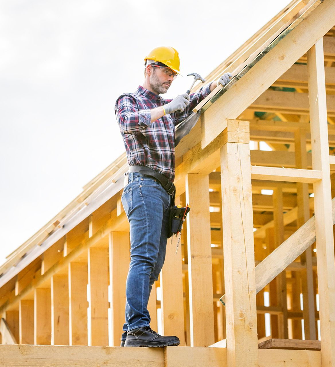 Construction worker in yellow hard hat standing on wooden roof frame and using a power tool