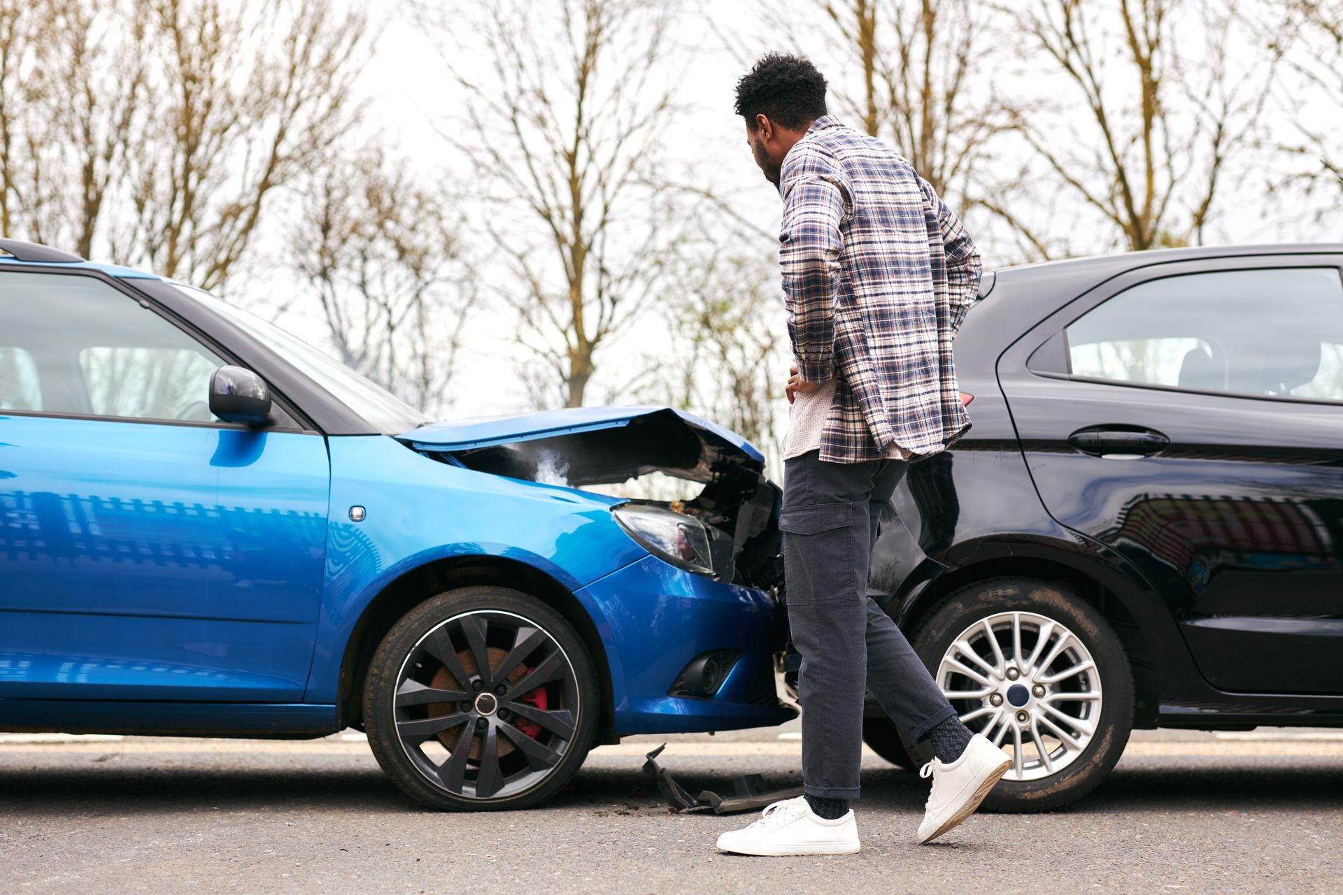 A person stands on the road looking at a blue car that has rear-ended a black car, causing hood damage.