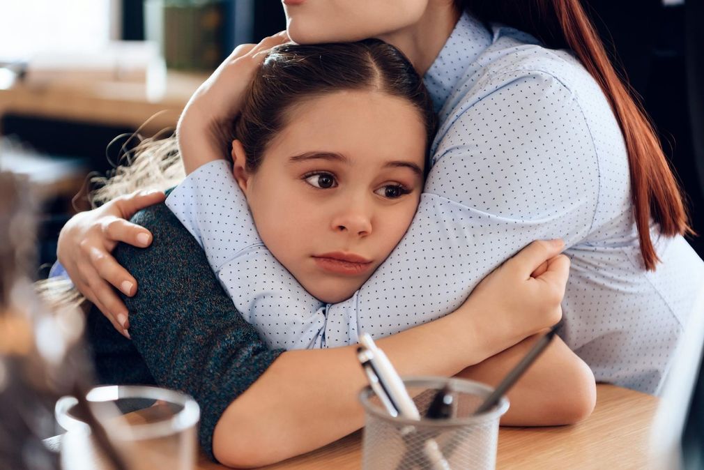 Child being comforted in a hug at a table, looking somber, with a pen cup in the foreground