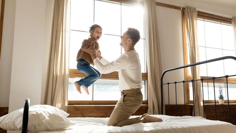 Adult lifting a smiling child on a bed in a sunlit bedroom near a window