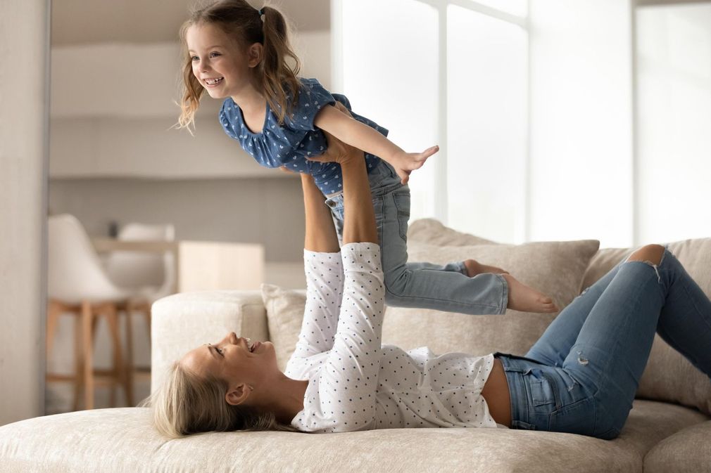 Woman and child play together on a couch in a bright living room, lifting the child into the air.