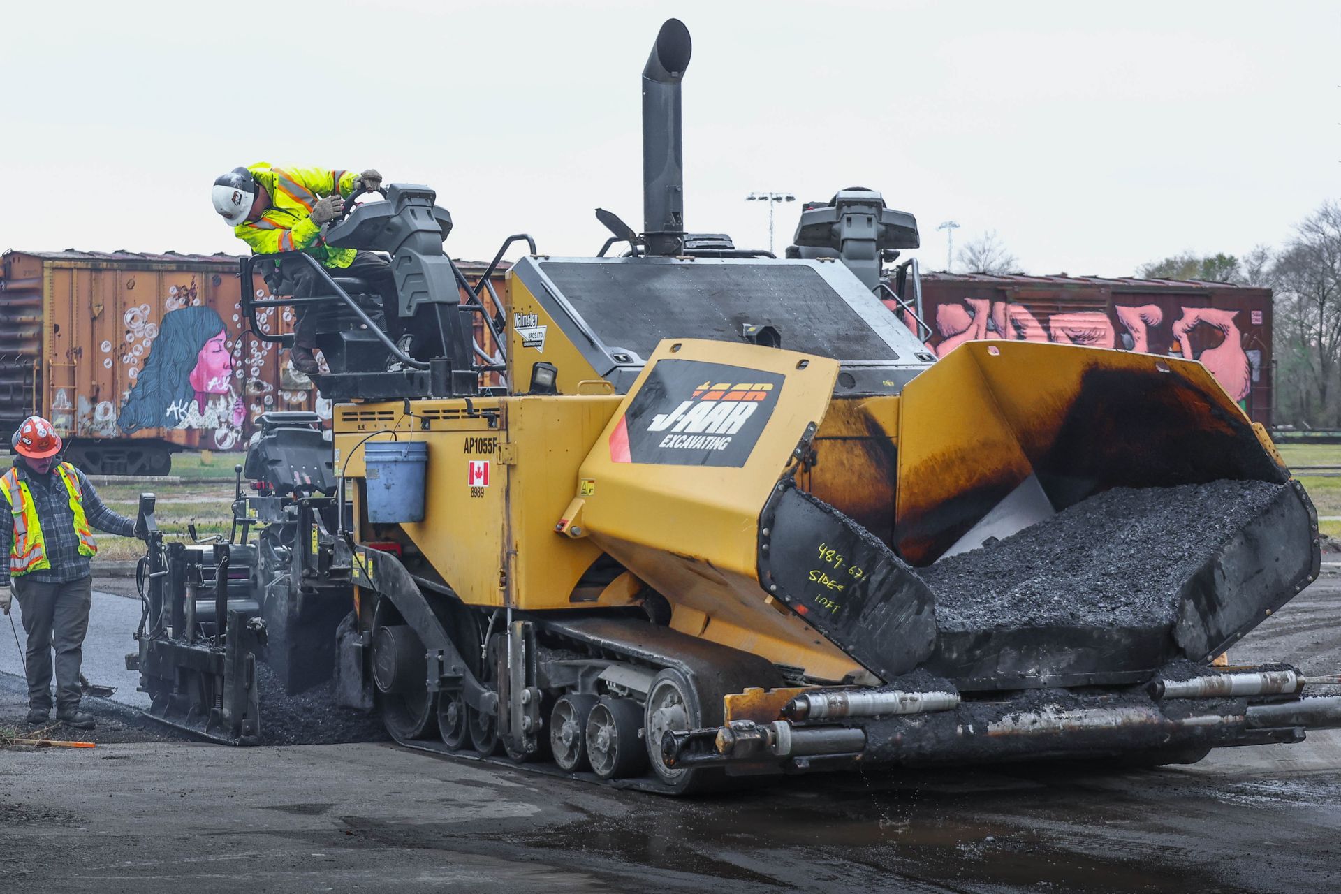 Workers on a yellow asphalt paver laying road surface on a wet construction site