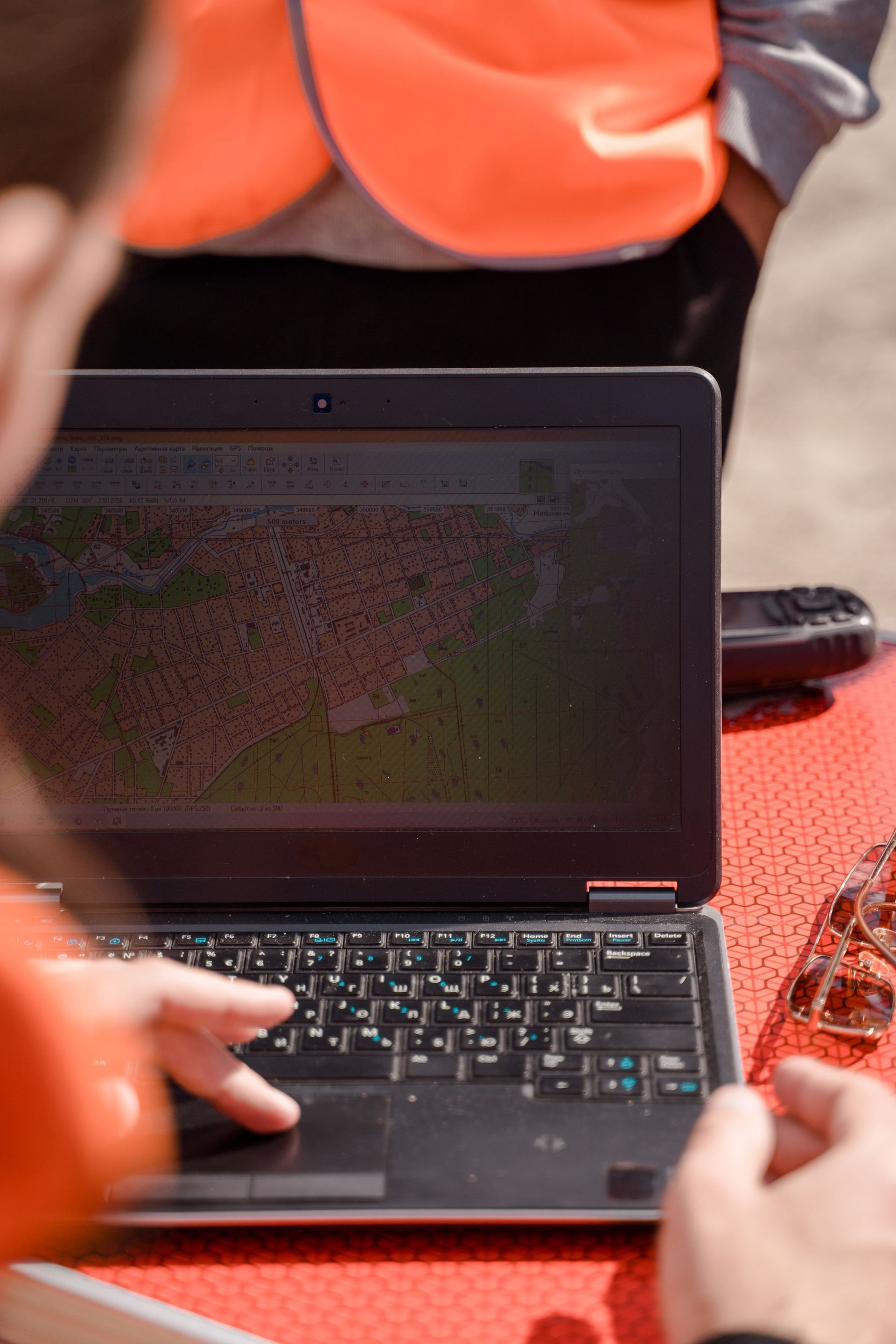 Person using a laptop outdoors at a table, with a bright orange vest visible nearby.