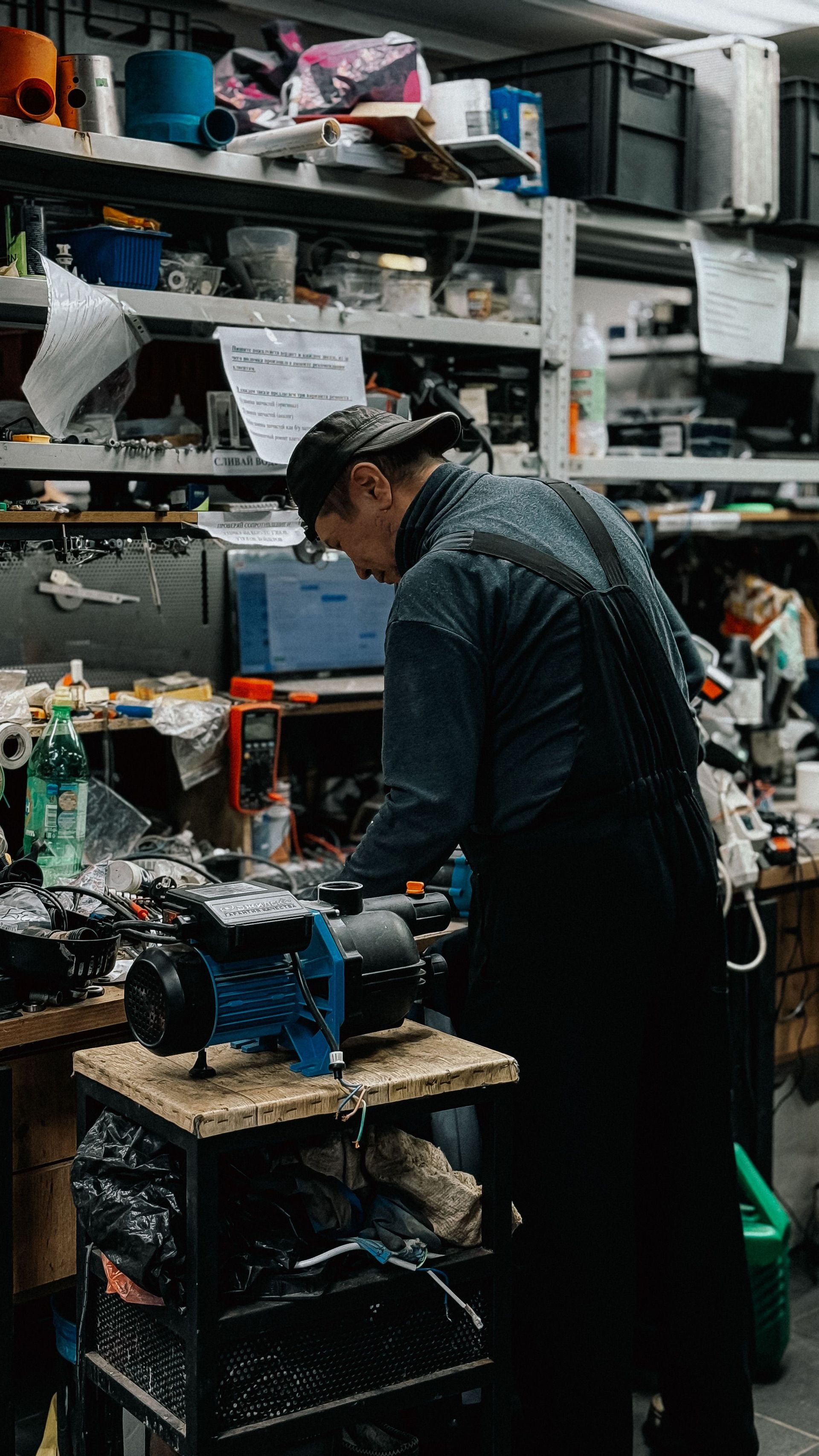 Person working at a cluttered workbench in a crowded workshop, surrounded by tools and equipment.