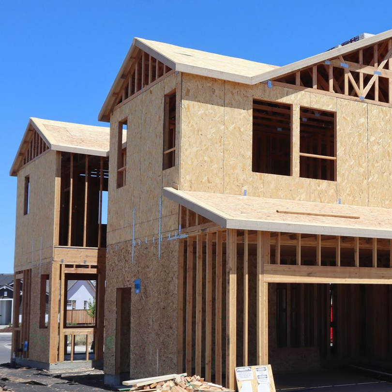 Wood-framed houses under construction with plywood siding and exposed roof trusses on a sunny day