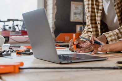 Person sketching with a pen beside a laptop on a desk with design tools and orange notebooks