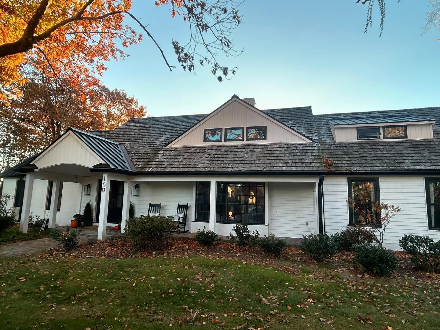 White house with dark roof and trim, porch with awning, front lawn with autumn leaves. New Roof