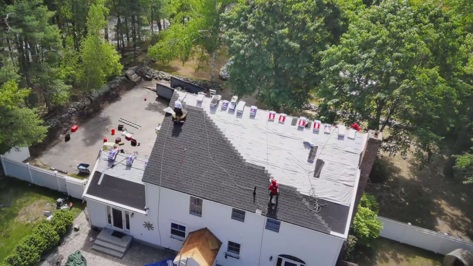 Workers on a roof with materials; white house, surrounding trees.