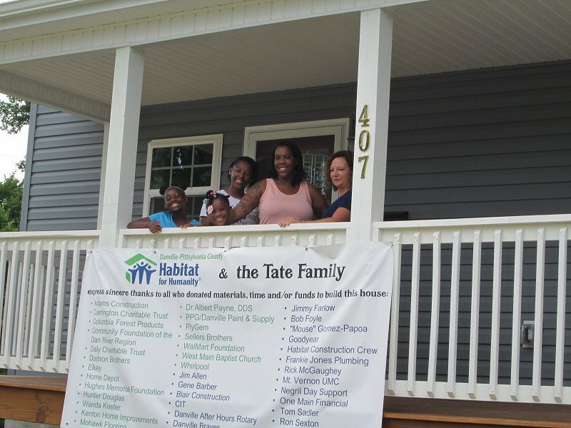 Three people on a front porch behind a Habitat for Humanity banner and house number 407
