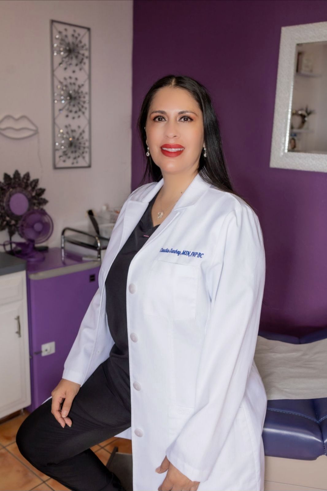A woman in a lab coat is sitting on a chair in front of a purple wall.