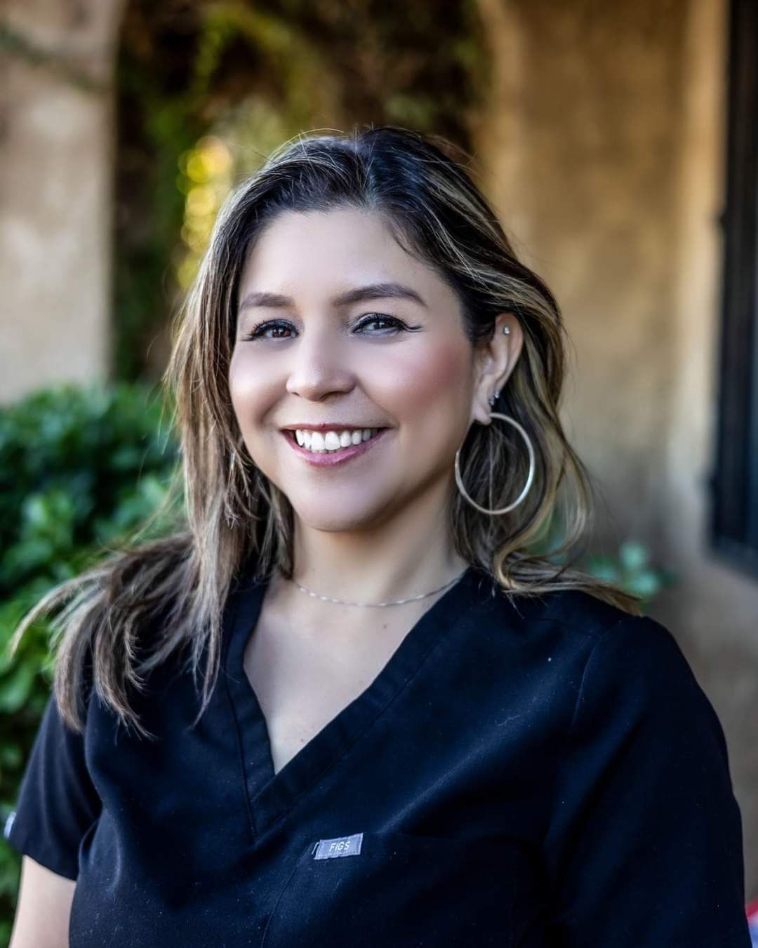 A woman wearing a black scrub top and hoop earrings is smiling for the camera.