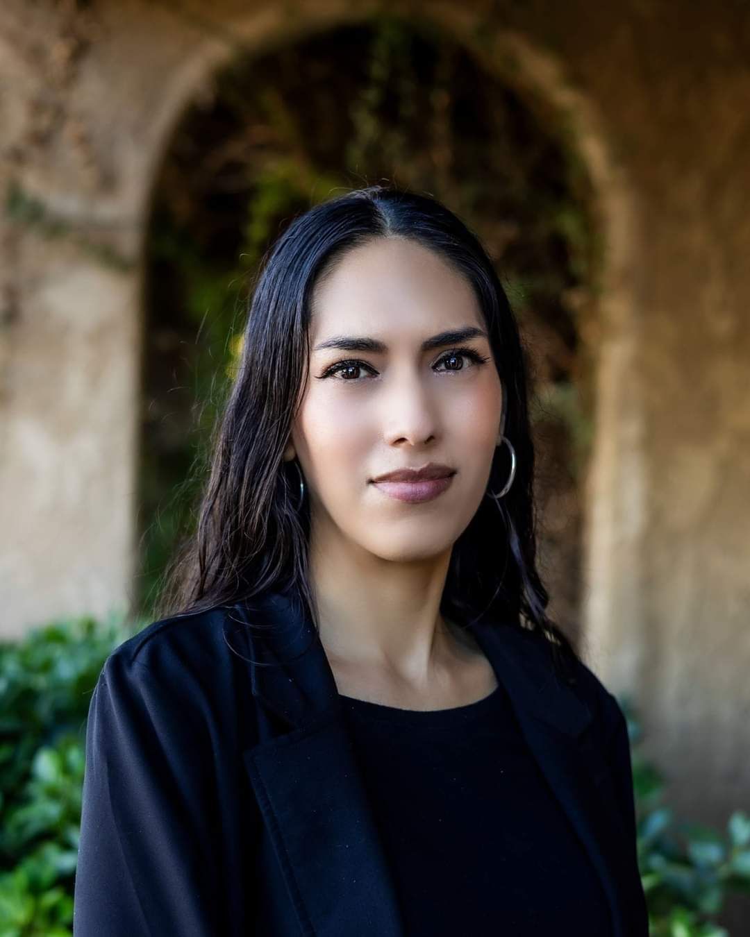 A woman in a black jacket and black dress is standing in front of a stone archway.