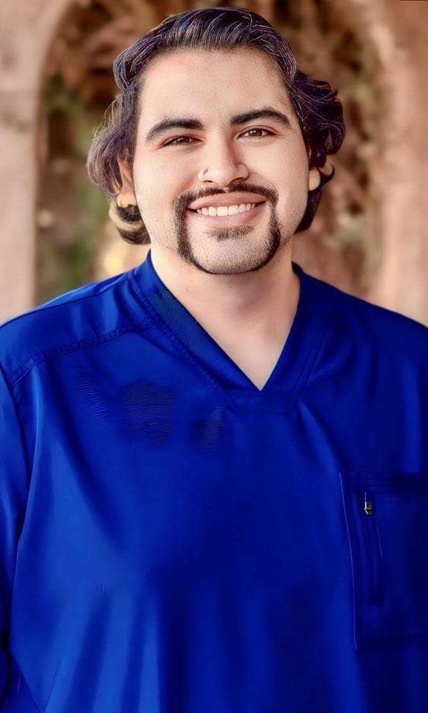 A man with a beard is wearing a blue scrub top and smiling.