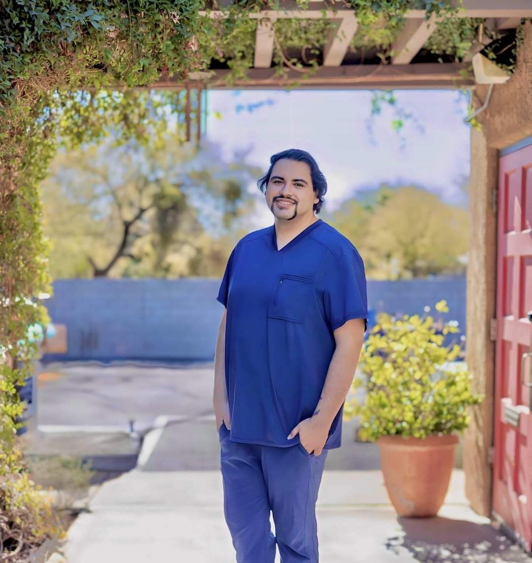 A man in a blue scrub top is standing in front of a red door.