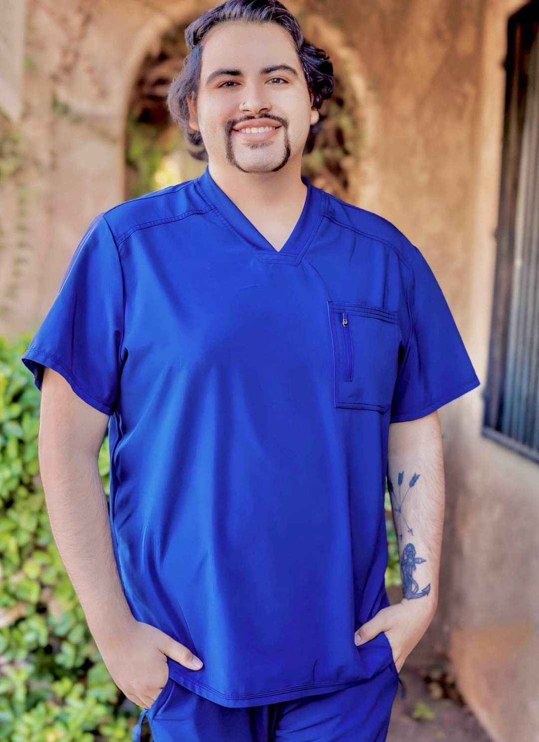 A man wearing a blue scrub top and pants is standing with his hands in his pockets.