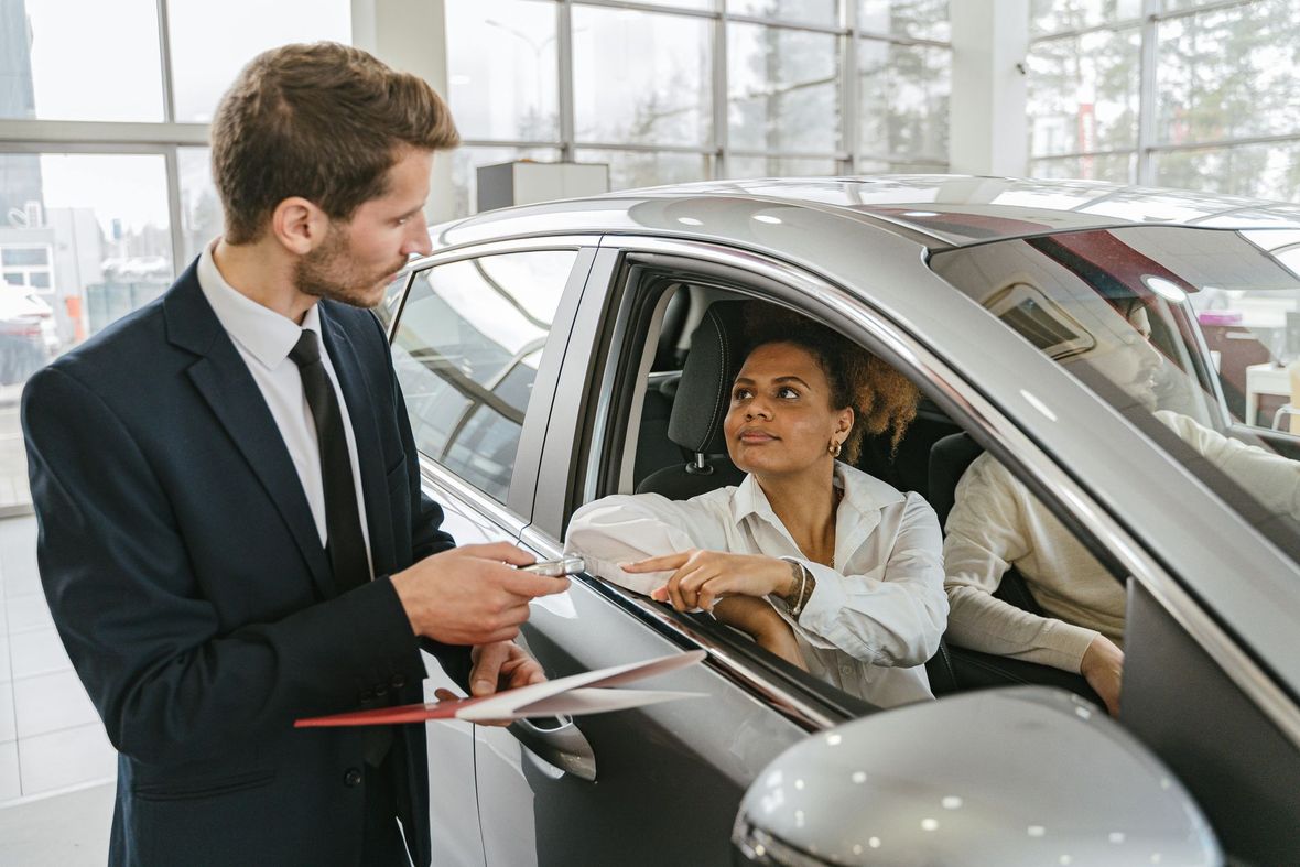 A salesperson in a suit holding a clipboard speaks to a customer seated in a silver car at a dealership.