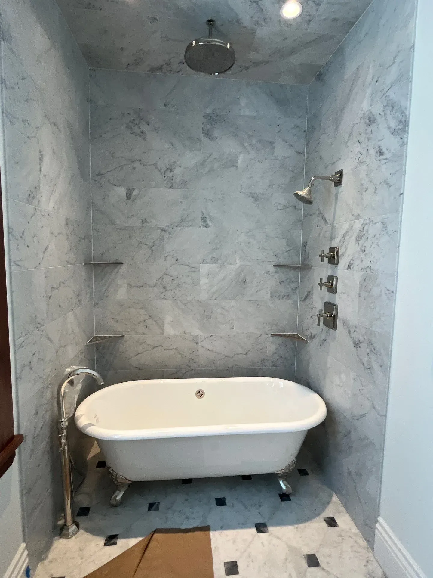 A clawfoot bathtub sits in a niche with grey-veined marble tile walls, a rainfall showerhead, and a hand shower faucet.