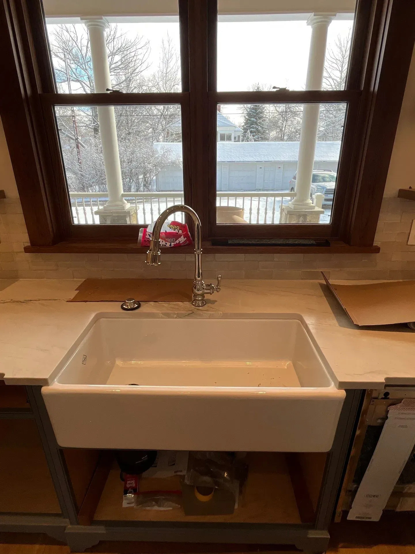 A white apron-front farmhouse sink with a chrome faucet set in a countertop in front of a window overlooking a snowy yard.