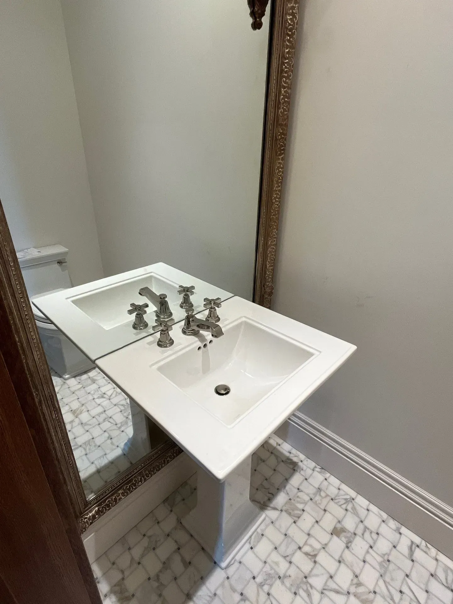 A pedestal sink with metallic fixtures stands in a bathroom with white, patterned marble tile flooring and a wall mirror.