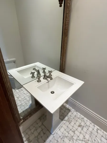 A pedestal sink with metallic fixtures stands in a bathroom with white, patterned marble tile flooring and a wall mirror.