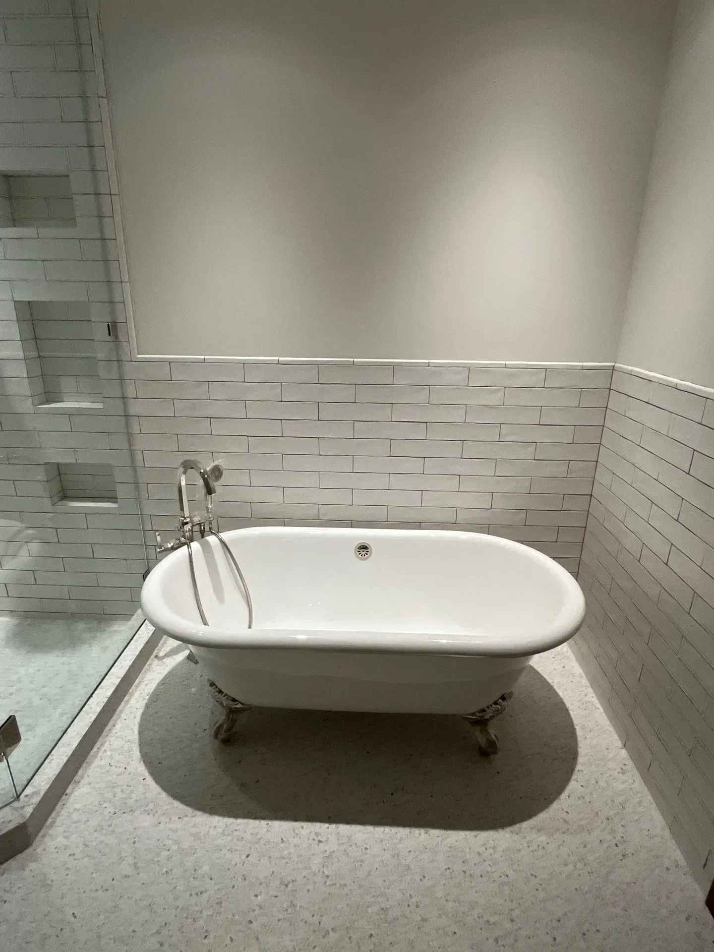 A white clawfoot bathtub sits in a modern bathroom with white subway-tiled walls and speckled terrazzo flooring.