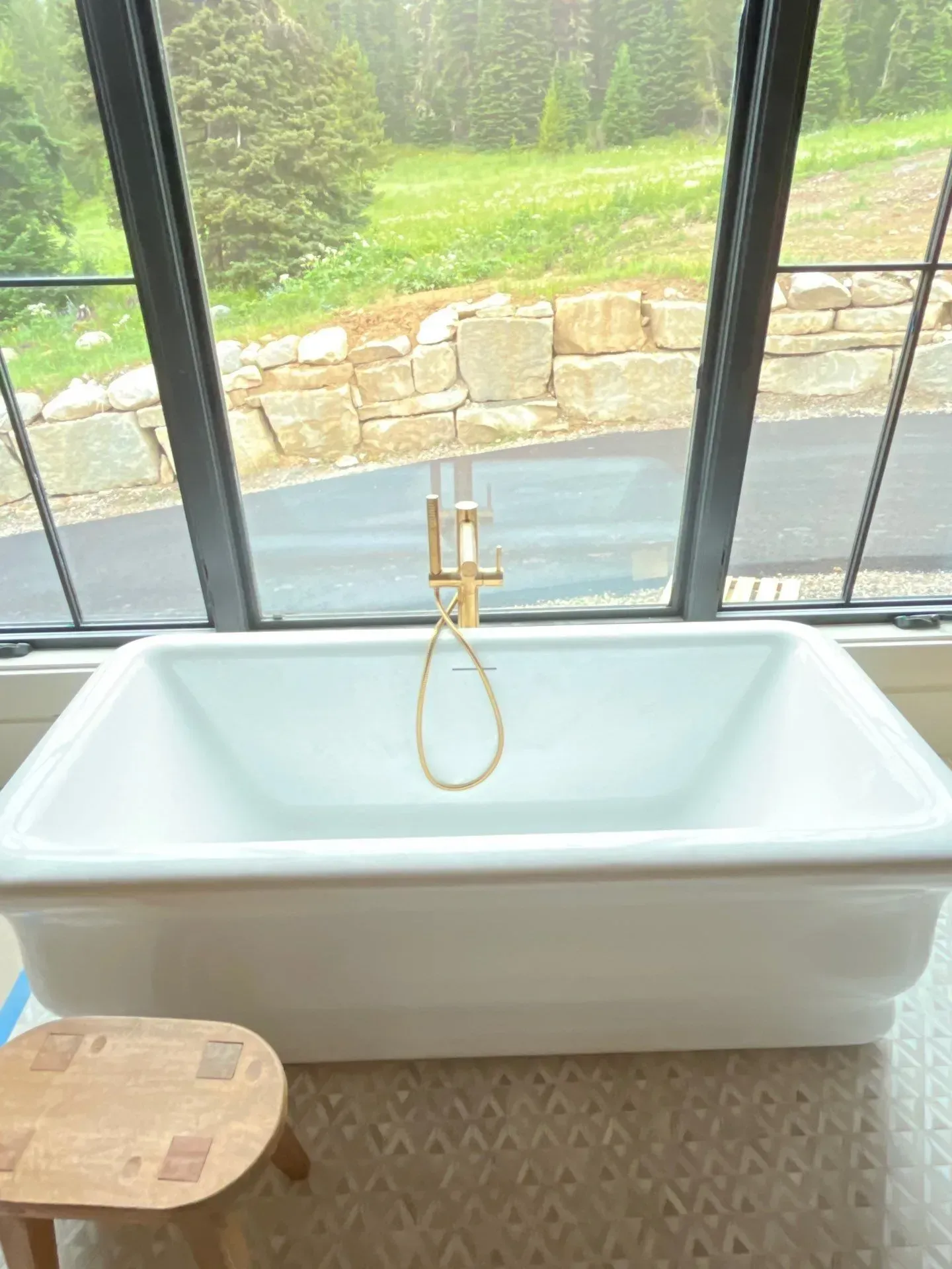 A white, rectangular soaking tub with gold fixtures stands before a large window overlooking a scenic, wooded outdoor area.