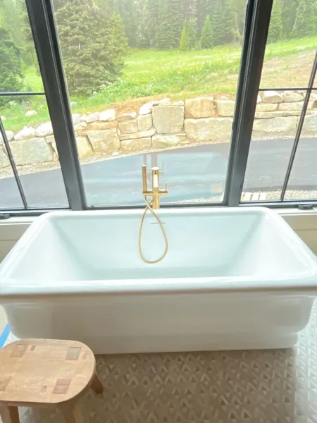 White rectangular bathtub with gold hardware in front of large windows overlooking a scenic, grassy outdoor landscape.