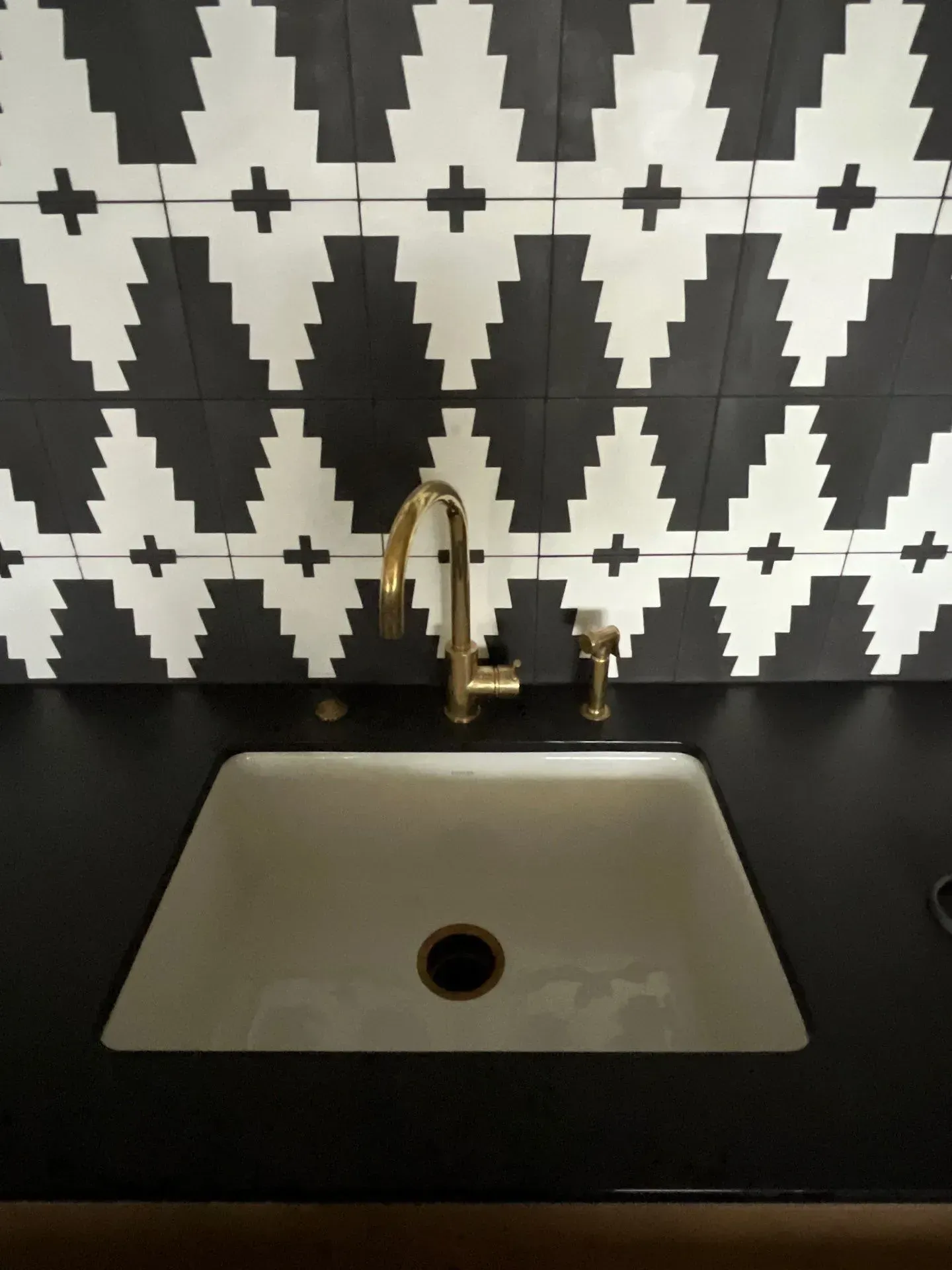 A white undermount sink with a gold faucet and side sprayer against a black countertop and bold geometric tile backsplash.