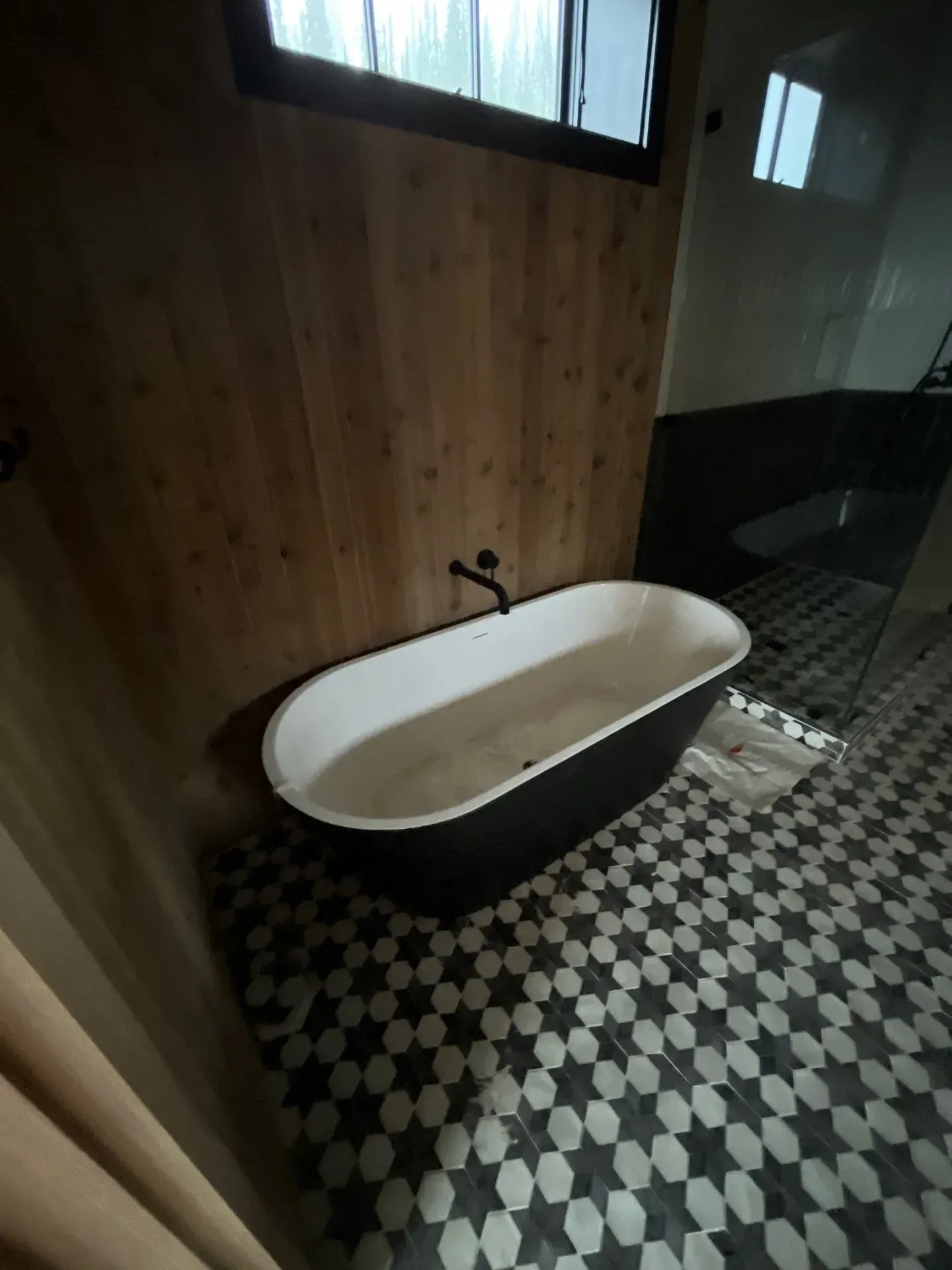 A black and white freestanding bathtub sits on a patterned tiled floor against a wood-paneled wall in a modern bathroom.