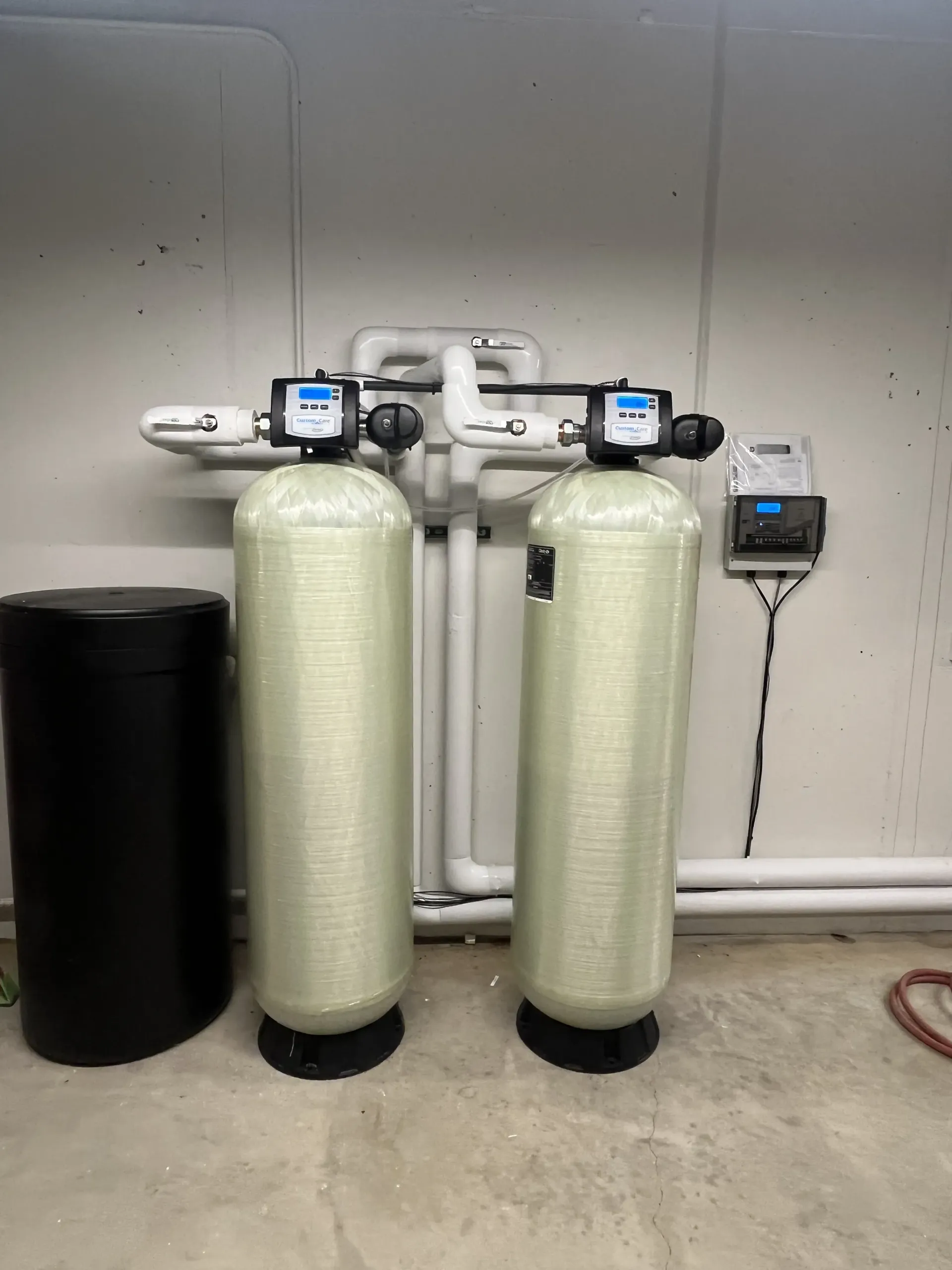Two tall water treatment tanks connected by white PVC pipes in a utility area next to a black brine tank.