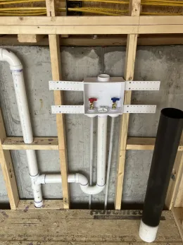 A washing machine outlet box with red and blue water valves installed in an unfinished wooden wall with PVC plumbing.