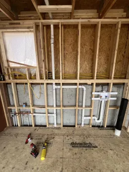 Interior view of unfinished residential framing featuring exposed wood studs, plumbing pipes, and a laundry box.