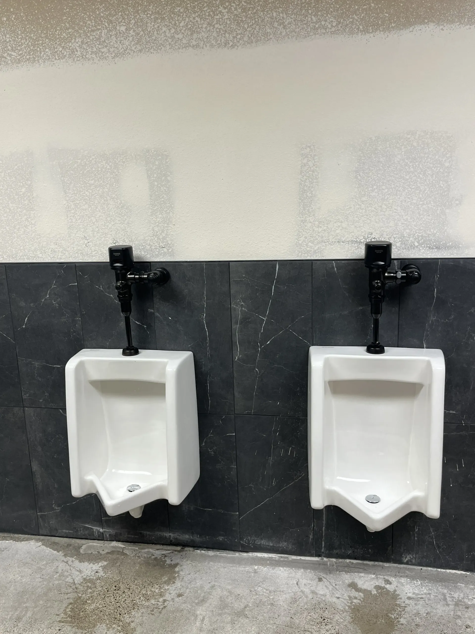 Two white wall-mounted urinals with black automatic flush valves against a dark tiled wall in a room under construction.