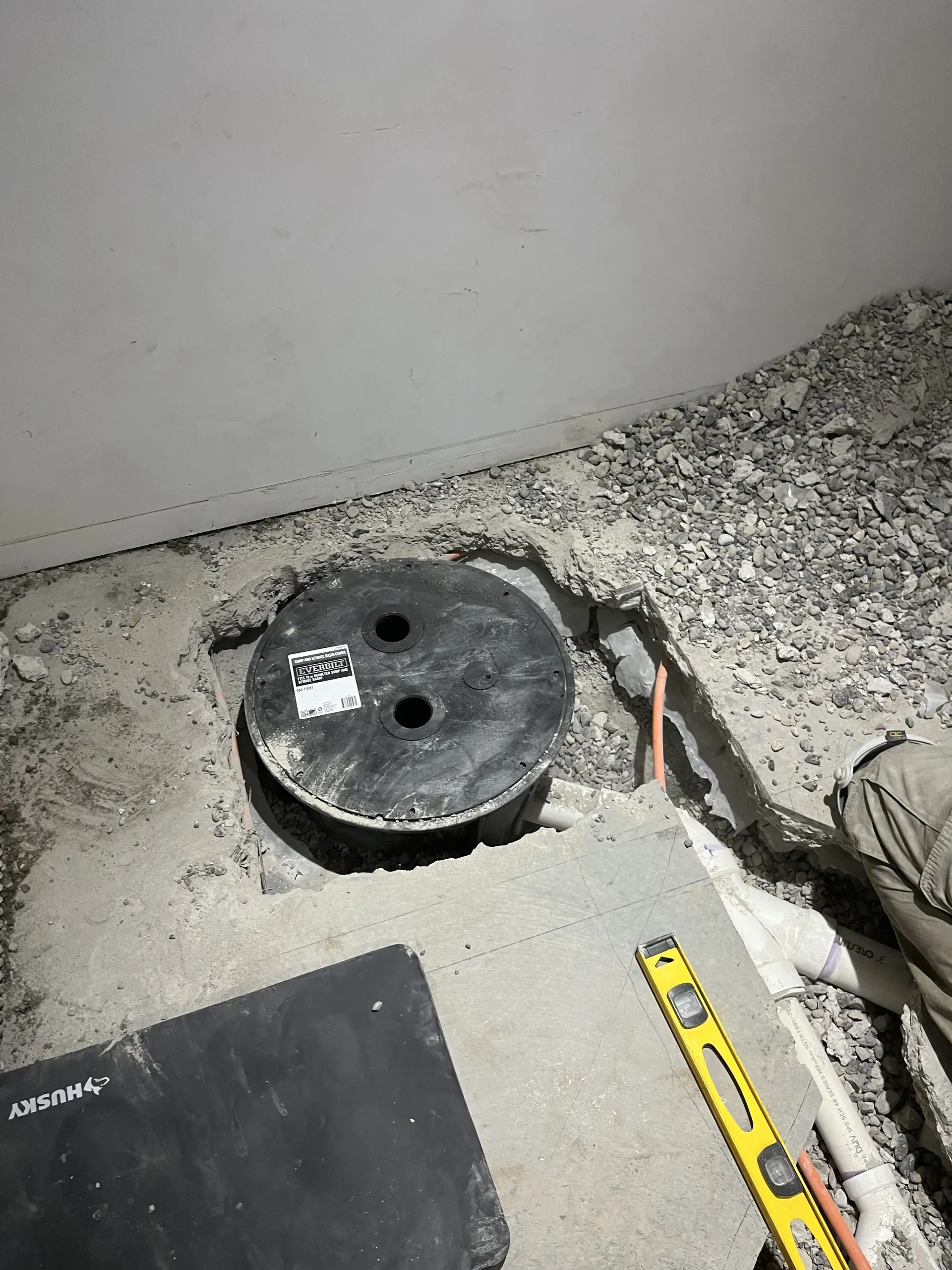 A black utility access lid sits in a concrete floor surrounded by gravel, with a yellow level tool nearby.