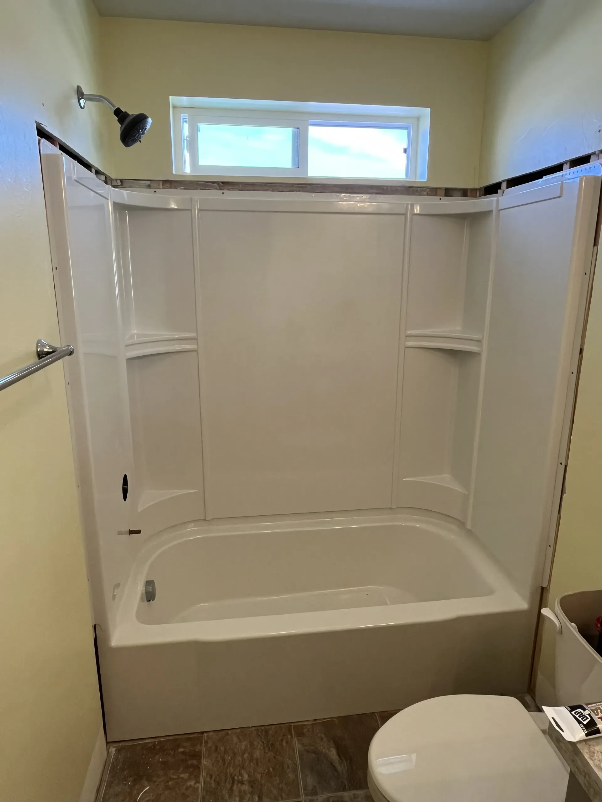 A white acrylic bathtub and shower surround in a bathroom with pale yellow walls and a window above.
