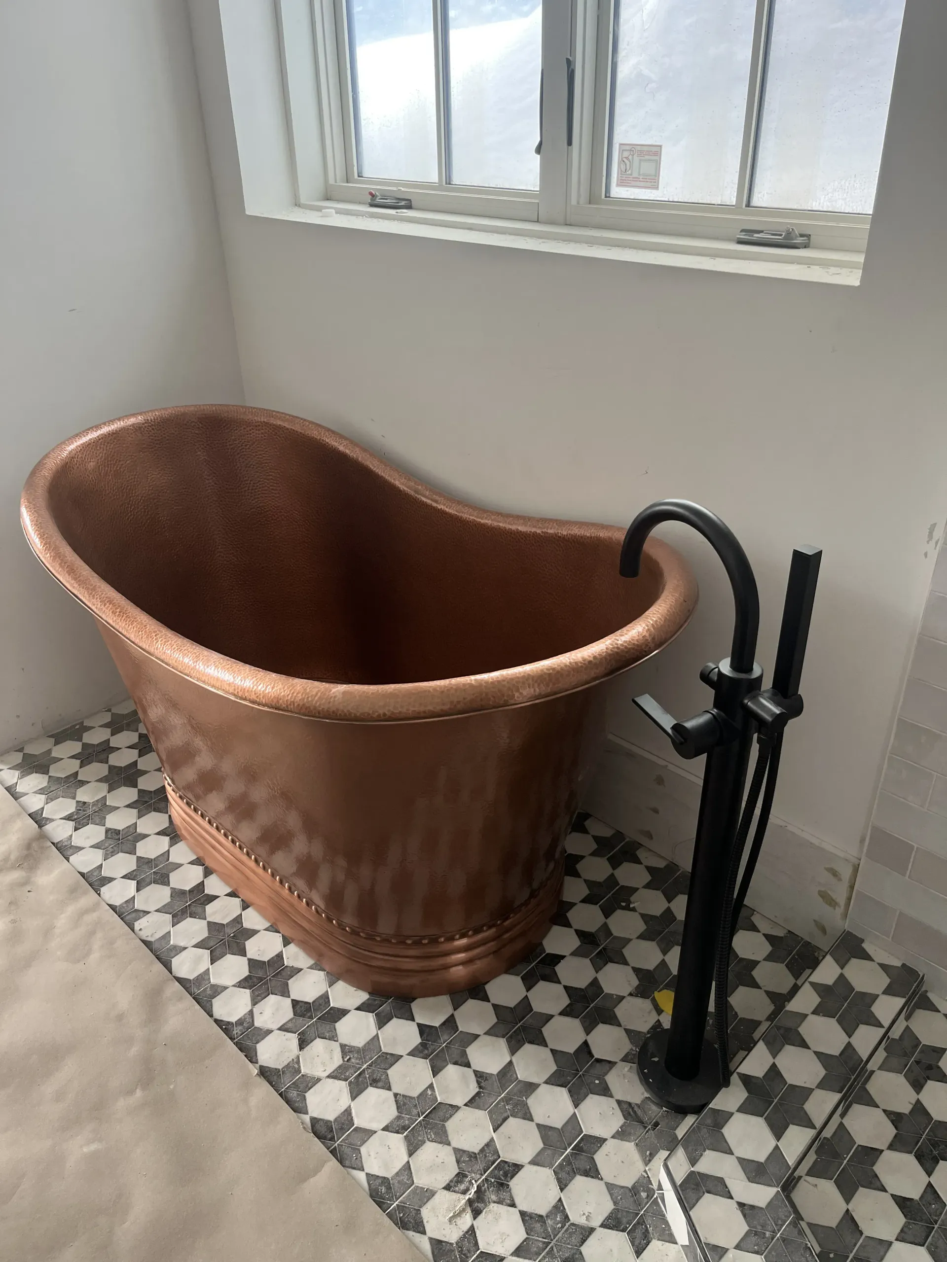 A copper, slipper-style freestanding bathtub sits on patterned tile flooring next to a matte black floor-mounted faucet.