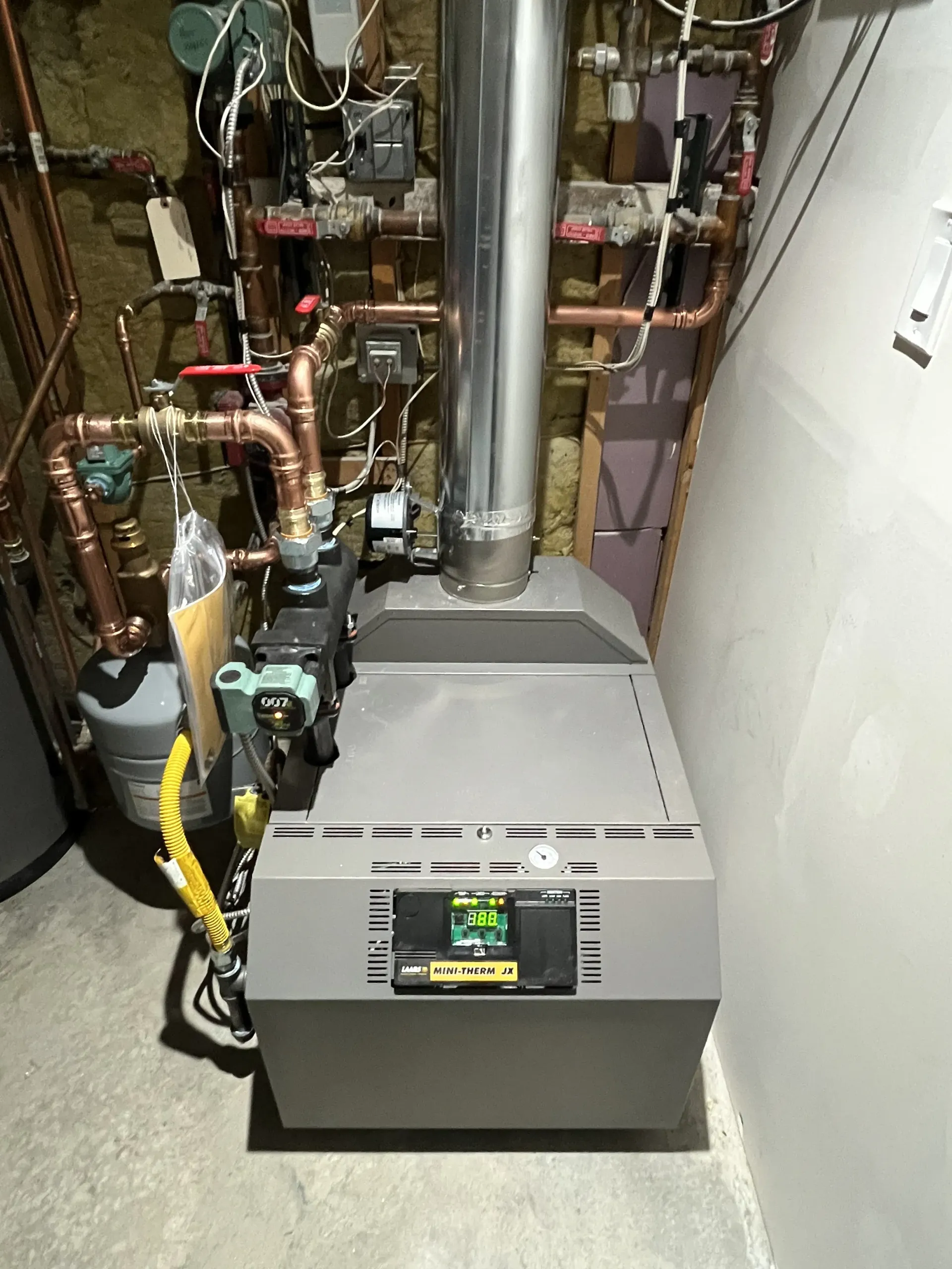 A gray home boiler unit with a silver exhaust vent, connected to copper piping and electrical wiring in a basement.