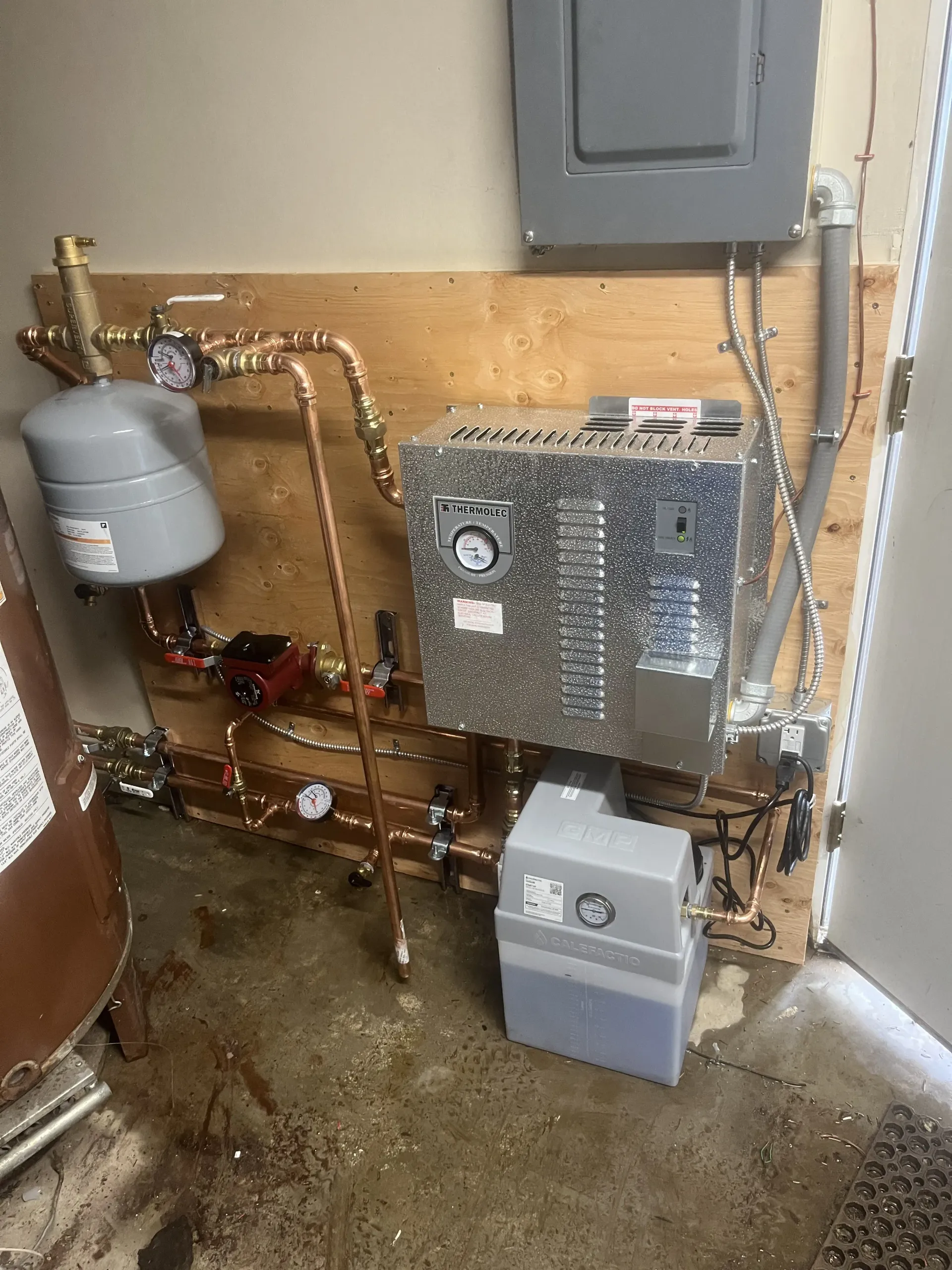 A residential heating system with a metal boiler unit, expansion tank, and piping mounted on a plywood wall.