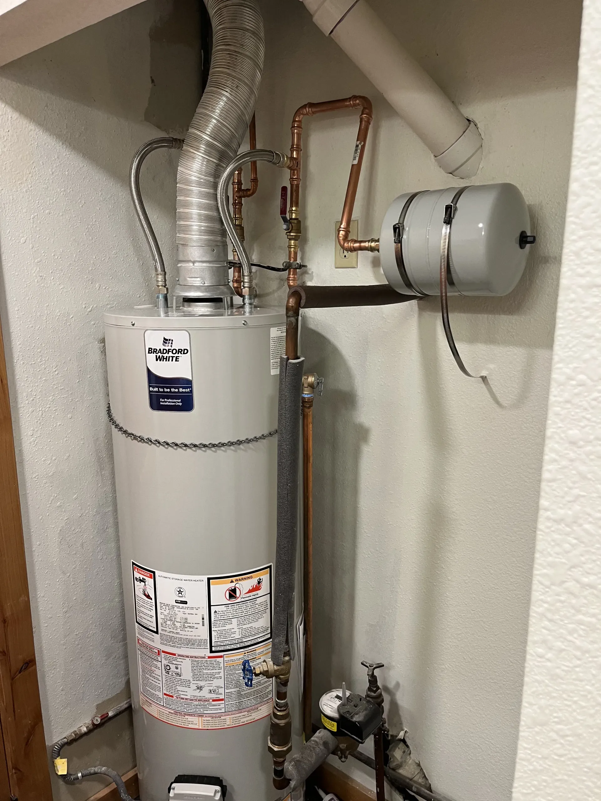 A grey residential water heater with a metallic exhaust vent and an attached expansion tank installed in a wall alcove.