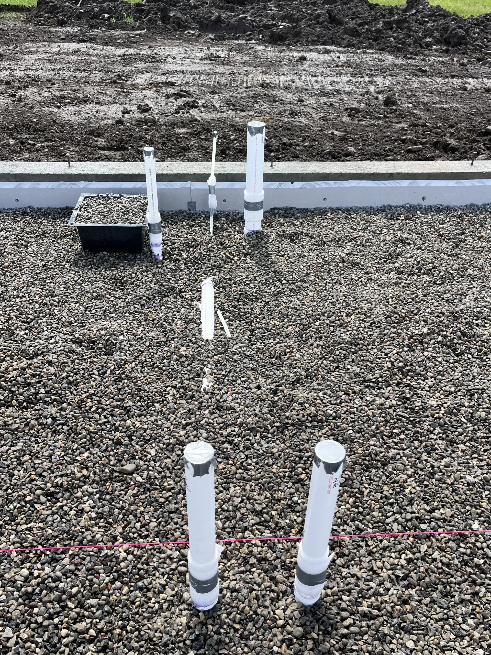 Several vertical white PVC pipes protrude from a gravel construction site, with a concrete curb in the background.