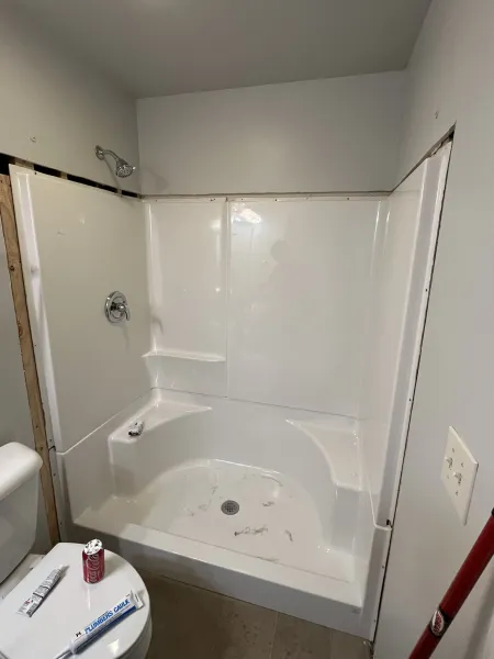 A newly installed white acrylic shower stall in a bathroom, featuring exposed wall edges and a nearby toilet.