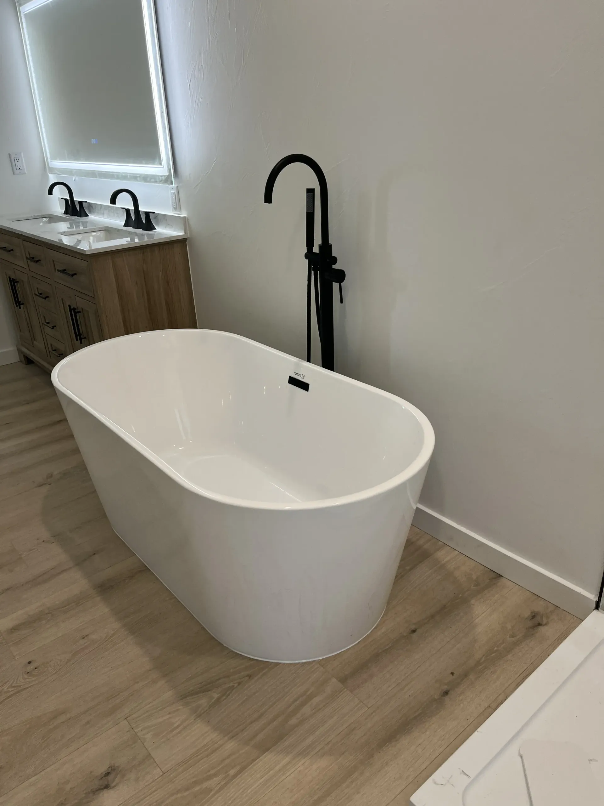 A modern white freestanding bathtub with a matte black faucet in a bright bathroom with light wood flooring.
