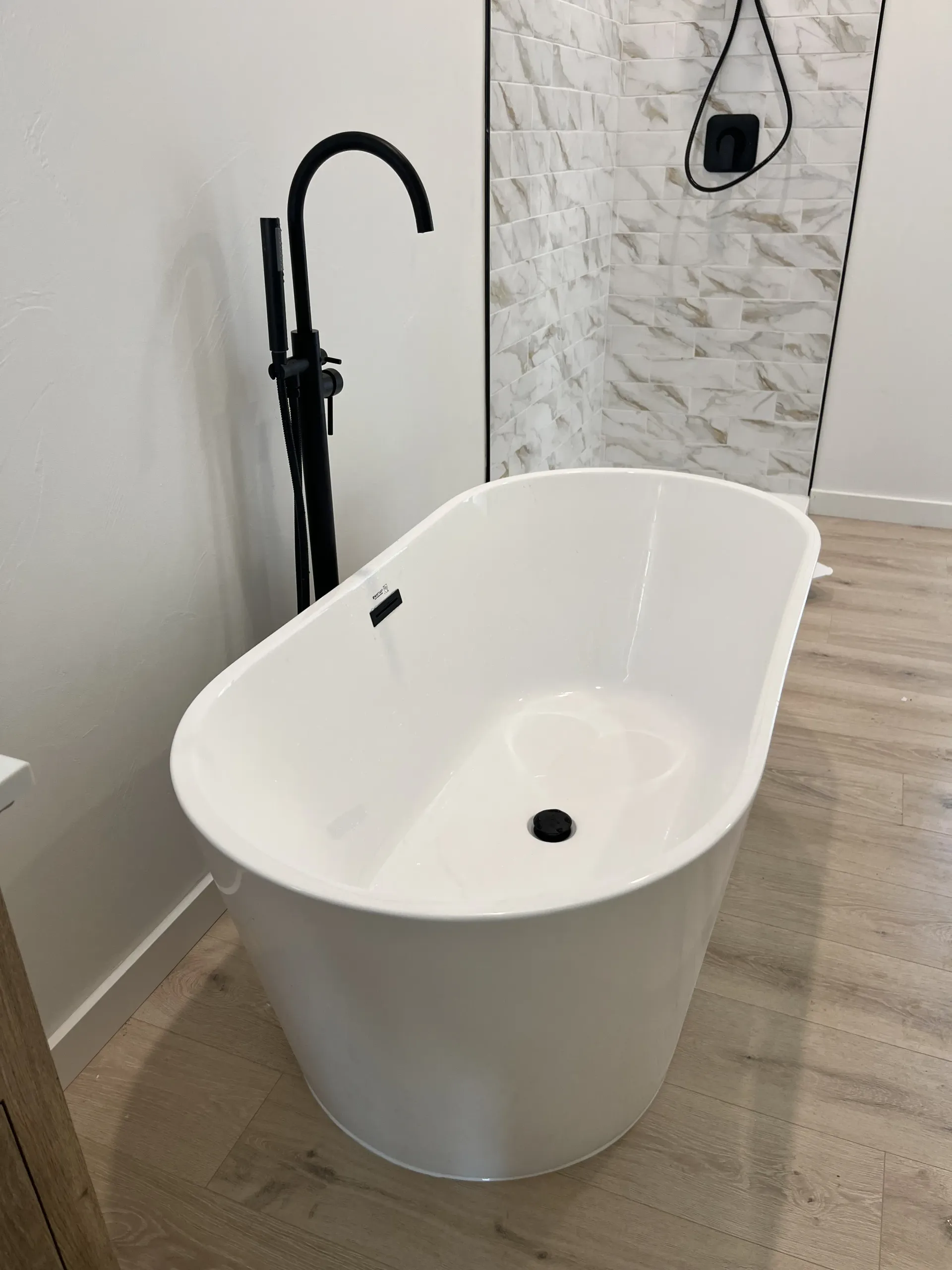 A white freestanding oval bathtub with a black floor-mounted faucet in a modern bathroom with light wood-look flooring.