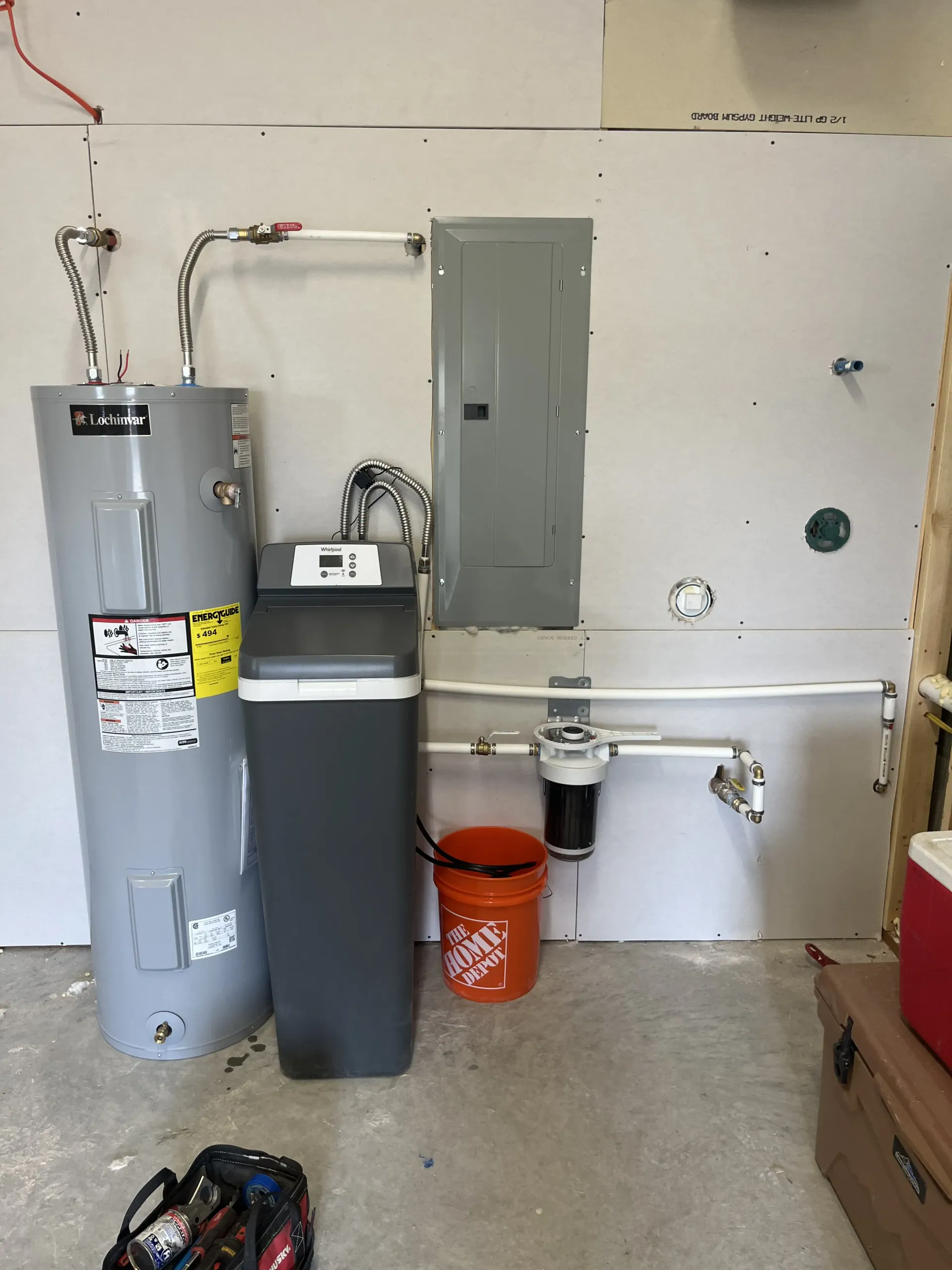 A water heater, softener, and filter system installed on a light-colored wall with a gray electrical panel in a garage.