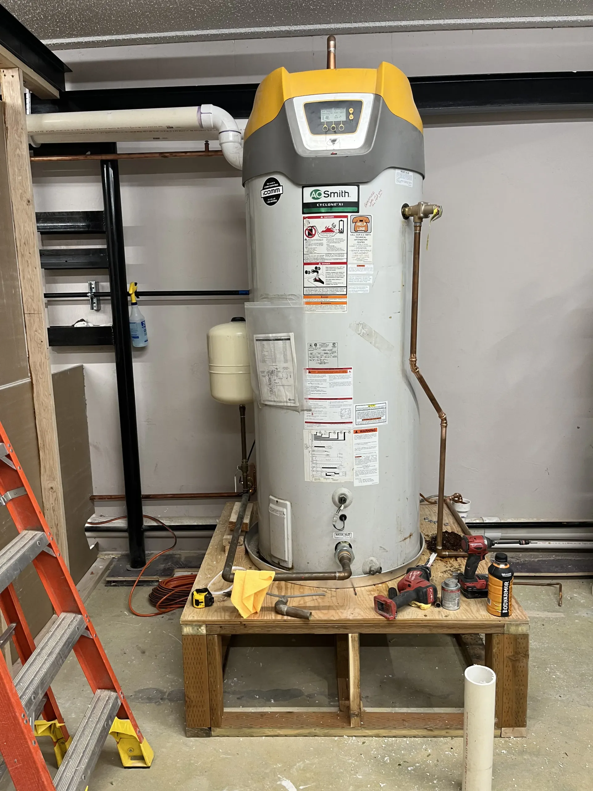A gray and yellow hybrid electric water heater sits on a wooden platform in a garage, with tools scattered nearby.
