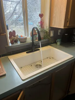A black kitchen faucet runs water into a white farmhouse sink with a metal grate at the bottom, placed under a window.