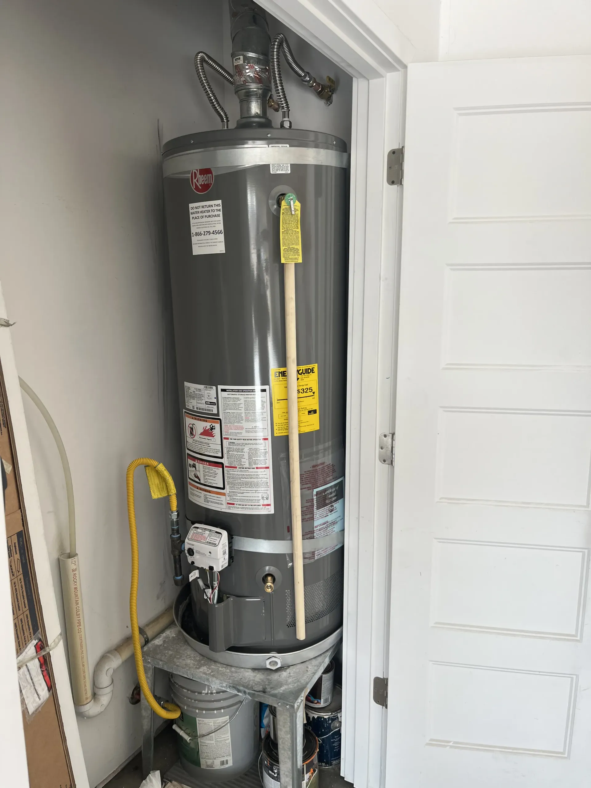 A grey water heater stands in a closet, resting on a stand inside a plastic bucket, with a yellow cord hanging nearby.