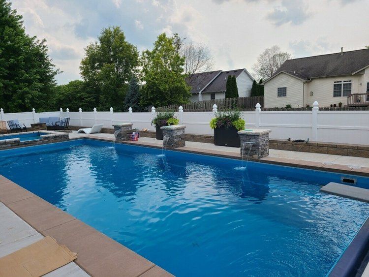 A swimming pool with blue water, surrounded by a concrete border and white fence, features stone pillars.